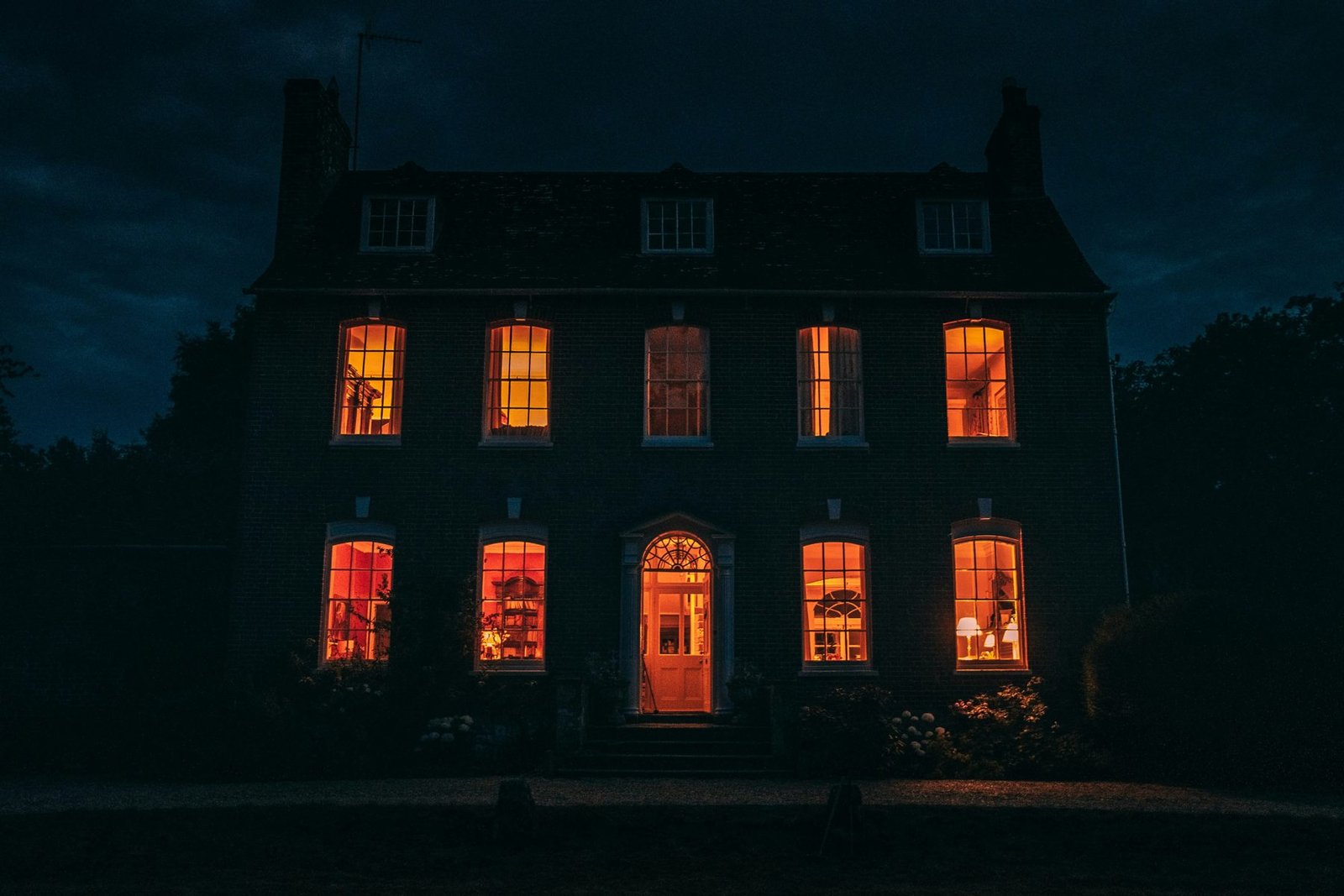 Large dark brick mansion at night with warm orange light glowing in every window, illustrative photo of the wealthy, secretive world of The Beast in Me